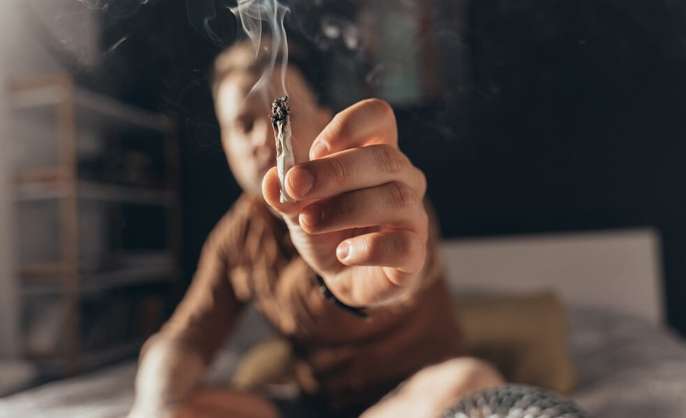 man offering a hand rolled cannabis cigarette while sitting at the bed
