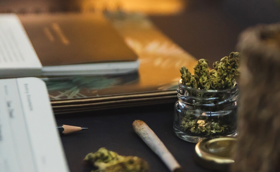 jar of cannabis buds, gold lid, notebooks, joint on black background
